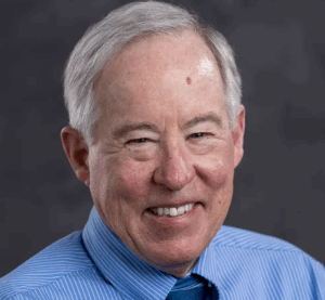 An older man with short gray hair smiles at the camera. He is wearing a blue striped shirt and matching tie, set against a dark, blurred background.
